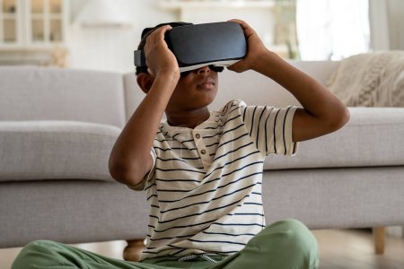 Téléchargez les photos : Curieux garçon afro-américain portant des lunettes 3D réalité virtuelle étude en ligne simulation monde virtuel à la maison. Petit écolier sérieux acquérir de l'expérience dans la connaissance de l'éducation avec l'aide VR lunettes - en image libre de droit