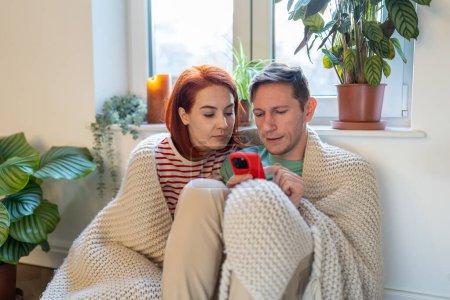 Photo for Family couple choosing products in online shop together on smartphone covered knitted plaid sitting near window in cozy interior. Man and woman having shopping, buying in internet scrolling pages. - Royalty Free Image