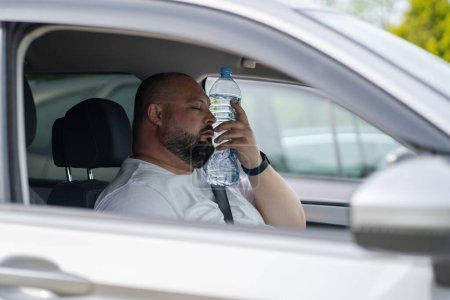 Exhausted overweight man driver suffering from heat inside car with broken air conditioner touch bottle of water to face closed eyes try cool. Male feel sick headache from hot air temperature 
