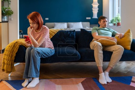 Couple man and woman ignoring each other. Wife looking at smartphone screen, tyrant husband sitting near on couch with evil face. Domestic violence, abuse, neurotic relationships, marital discord.