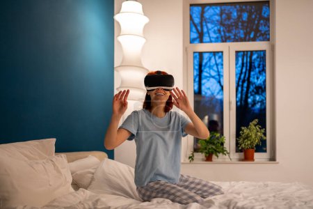 Foto de Mujer usando gafas de realidad virtual en la cama en casa. Chica juega videojuegos a través de un dispositivo especial, simulador de casco. Mujer feliz con gafas especiales para juegos de realidad virtual explora el mundo no real - Imagen libre de derechos