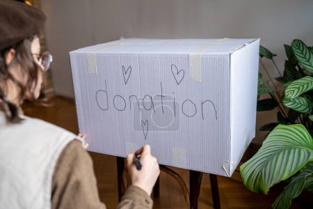 Téléchargez les photos : Millennial girl écrit un don de mots sur une boîte en carton tout en triant les vêtements usagés pour faire un don à la maison, femme faisant un don de vêtements indésirables à un organisme de bienfaisance, aidant les personnes dans le besoin - en image libre de droit