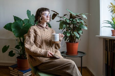 Foto de Chica milenaria pensativa con gafas sentadas con teléfono y taza de té en la acogedora casa de la selva urbana, mujer soñadora reflexiva se sienta contra plantas de interior en macetas verdes mirando por la ventana y reflexionando - Imagen libre de derechos