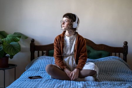 Téléchargez les photos : Adolescente détendue portant des écouteurs assis sur le lit en pose de lotus et regardant de côté, écouter du contenu audio, apprendre la langue avec des podcasts. Millennial femme écouter de la musique dans les loisirs - en image libre de droit