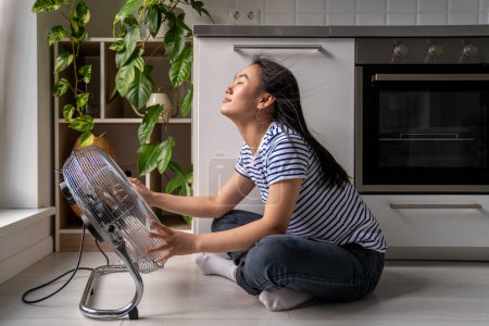 Téléchargez les photos : Cheerful Asian girl enjoys cold wind from electric fan sits on floor in kitchen. Young Japanese woman resting at home and sits by fan and enjoys the cool breeze. Summertime, heat concept. - en image libre de droit