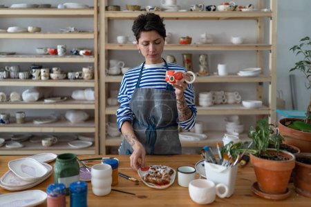 Photo for Woman ceramist having morning snack in ceramics workshop, drinking tea with delicious freshly baked bun at workplace in pottery studio, producing handmade pottery. Female potter taking break - Royalty Free Image