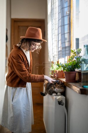 Photo for Love your pet. Smiling millennial girl petting furry cat to relieve stress while standing near window at home, owning companion animal. Companion animals, benefits of pet ownership - Royalty Free Image
