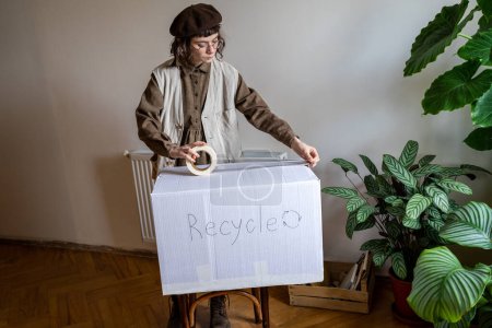 Foto de Hipster chica pegando cinta adhesiva en la caja de cartón mientras clasifica la ropa no deseada para el reciclaje en casa, enviando ropa usada vieja para el reciclaje textil. Reducir y reutilizar - Imagen libre de derechos