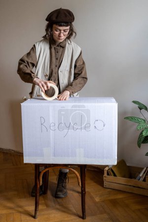 Foto de Hipster niña de pie en casa sosteniendo cinta adhesiva embalaje de ropa vieja, zapatos y textiles en la caja de cartón para el reciclaje, la limpieza de armario, el envío de ropa al centro de reciclaje. Ecológico hogar - Imagen libre de derechos