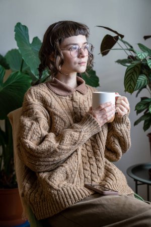 Téléchargez les photos : Adolescente songeuse avec une tasse de thé à la maison, pensant ou rêvant, assise avec une boisson chaude dans un intérieur confortable de jungle urbaine et regardant par la fenêtre. Concept de rêverie - en image libre de droit