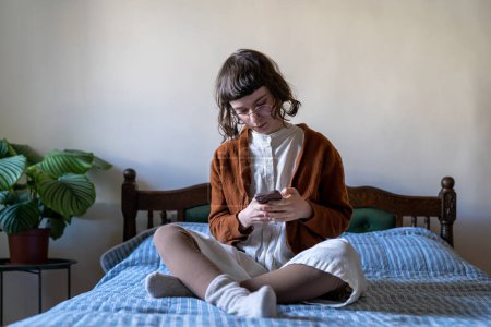 Photo for Pensive dreamy student girl sitting crosslegged on bed looking to smart phone screen, reading online book, chatting, browsing social networks, relaxing home after college. Internet addiction - Royalty Free Image