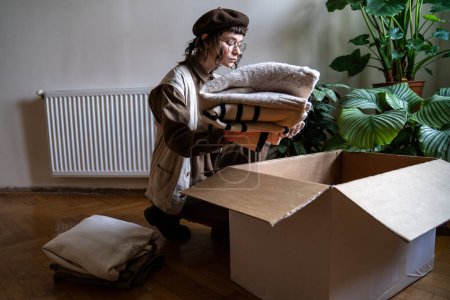 Photo for Clothing donation. Caring volunteer woman in eyeglasses puts used clothes in cardboard box. Packing warm sweaters for recycling, conscious consumption things for flea market. Problem waste in fashion - Royalty Free Image