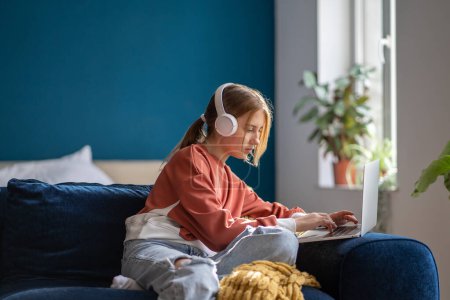 Téléchargez les photos : Une adolescente concentrée portant un casque tapant du texte sur un ordinateur portable détendue assise sur un canapé dans le salon. Pensive adolescent écolier étudiant en ligne, enseignement à distance, en utilisant l'ordinateur à la maison. - en image libre de droit