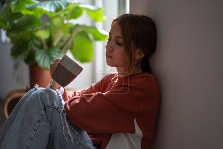 Photo for Frustrated sad teenage girl sitting on floor with cup devastated thinking about trouble, broken heart. Young woman have bad mood due to hormones in period. Depressive thoughts from problems at school. - Royalty Free Image