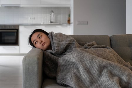 Téléchargez les photos : Inintéressé fatigué asiatique coréen jeune femme regarde tristement loin de mauvaise humeur se trouve sur le canapé sous couverture de laine à la maison pense à des problèmes de dépression dans la santé mentale, burnout ou le cœur brisé - en image libre de droit