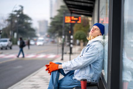 Photo for Tired guy tourist with coffee take away waiting bus. Blonde hair millennial man on bus stop in city. Fair-haired teen student waiting transport very long time in morning - Royalty Free Image