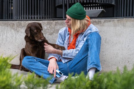 Téléchargez les photos : Hipster gars et chien de compagnie mignon sans abri assis ensemble à l'extérieur, jeune homme parlant avec un animal errant amical. Concept de relations homme-animal - en image libre de droit