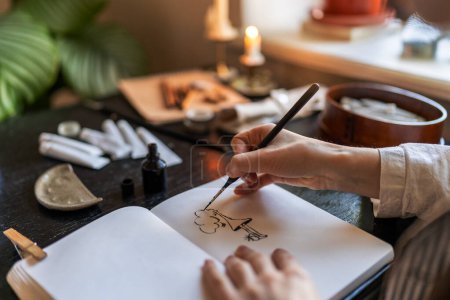 Photo for Close up of artist holding fountain pen drawing with ink while sitting at table with sketchbook and painting tools, drawing at home, creating artwork. Creative people and inspiration concept - Royalty Free Image