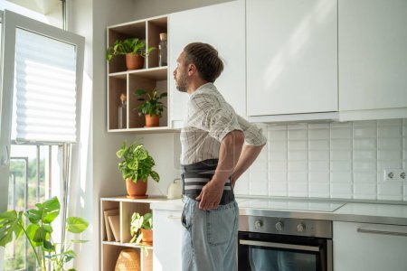 Man in sick leave at home on kitchen wearing back support belt corset on lower back during exacerbation to treatment of hernia, postoperative recovery. Back pain, spine health problems concept. 