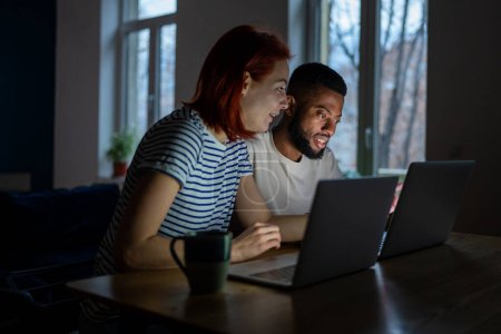 Photo for Happy diverse entrepreneurial couple have small online business at home. Multiracial freelancers man and woman sitting at table working on laptops together in late evening. Family entrepreneurship. - Royalty Free Image