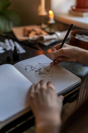 Photo for Closeup of artist holding fountain pen drawing with ink while sitting at table with sketchbook and painting tools, drawing at home, creating artwork. Creative people and inspiration concept - Royalty Free Image