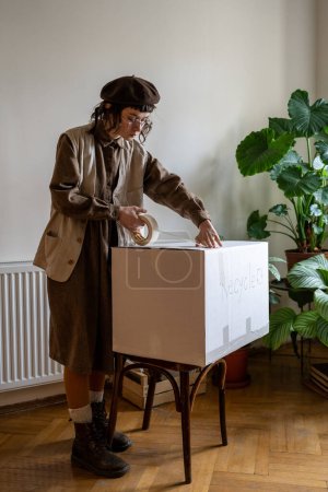 Téléchargez les photos : Hipster girl debout à la maison tenant du ruban scotch emballant de vieux vêtements, chaussures et textiles dans une boîte en carton pour le recyclage, le nettoyage de l'armoire, l'envoi de vêtements au centre de recyclage. Maison écologique - en image libre de droit
