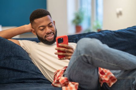 Foto de Relajado afroamericano sonriente hombre se sienta en el sofá mira el teléfono inteligente en casa en la sala de estar. Chico negro interesado feliz riéndose de vídeo divertido o broma, charlando con amigos en las redes sociales. - Imagen libre de derechos
