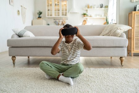 Foto de Niño afroamericano sentado en el suelo en la sala de estar con auriculares de realidad virtual que exploran el mundo virtual. Niño escolar que usa la tecnología VR para aprender. Realidad aumentada para niños - Imagen libre de derechos