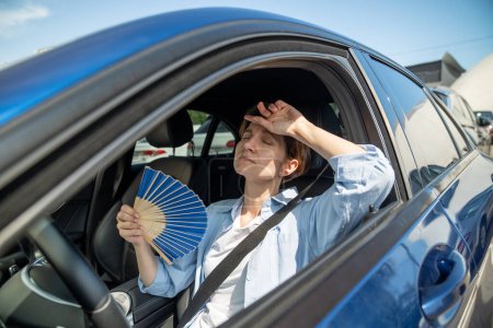 Photo for Exhausted tired middle aged woman drives car waves blue fan suffers from stuffiness stands in urban traffic jam in summer hot weather. Overheating, high temperature in car with broken air conditioner. - Royalty Free Image