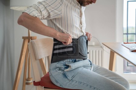 Photo for Man sitting on chair wearing back brace to reduce hernias, using decompression corset for herniated discs at home, relieving symptoms of degenerative disc disorder. Recovery after lower back surgery - Royalty Free Image