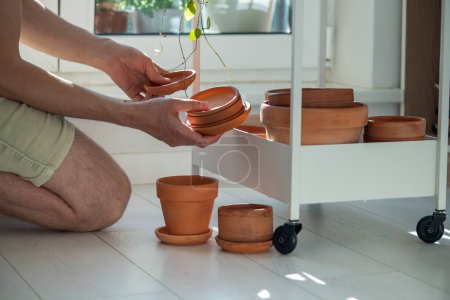 Man gardener holding tray for terracotta pot for transplanting plant at home sitting on floor and choose flowerpot of right size and diameter. Indoor gardening, plant lovers concept. 
