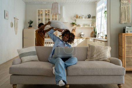 Photo for Displeased African American mother freelancer sitting on sofa at home working on laptop while small child distracts and hits mom pillow. Hyperactive boy asks for attention from parent - Royalty Free Image