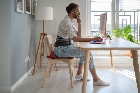 Photo for Focused male freelancer wearing lumbar brace sitting at table using computer, young man use orthopedic corset belt while working remotely with herniated disc. Work from home and back pain - Royalty Free Image