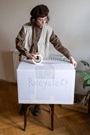 Foto de Hipster chica pegando cinta adhesiva en la caja de cartón mientras clasifica la ropa no deseada para el reciclaje en casa, enviando ropa usada vieja para el reciclaje textil. Reducir y reutilizar - Imagen libre de derechos