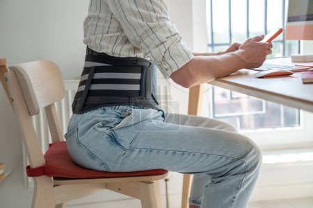 Photo for Man freelancer wearing medical orthopedic corset sitting at table working remotely from home, remote worker using lower back brace sits in front of computer, treating chronic back pain - Royalty Free Image