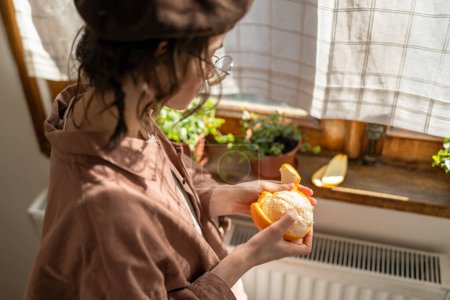 Photo for Stylish girl in beret peels orange near windowsill with houseplants at home. Sunny day in harmony with yourself. Artist woman holding fresh fruit in hand enjoy fragrance pondering new idea for painted - Royalty Free Image