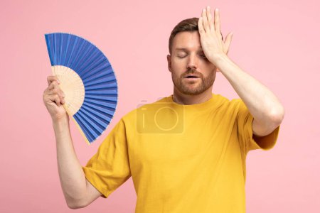 Téléchargez les photos : Sweaty fatigué homme d'âge moyen toucher le front en utilisant un ventilateur en papier souffrent de chaleur, se sent lent. Homme mécontent se refroidissant par temps chaud d'été, isolé sur fond rose studio. Concept de surchauffe - en image libre de droit