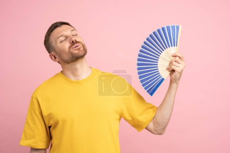 Photo for Overheated man using paper fan suffer from heat, sweating, cools herself due problem no air conditioner at home at summer weather, enjoying fresh air and closed eyes isolated on studio pink background - Royalty Free Image