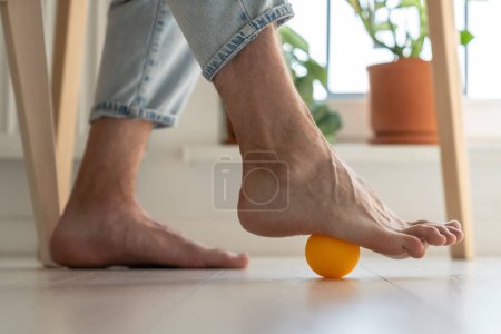 Téléchargez les photos : Homme utilisant la boule de silicone pour le massage des pieds pendant le long travail sédentaire, jambes gros plan. Exercices physiques pour la récupération des pieds après traumatisme, amélioration de la circulation sanguine, prévention des maladies des jambes. - en image libre de droit