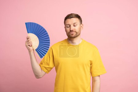 Photo for Overheated man waving paper fan suffer from heat, feels sluggish and drowsiness. Unhappy exhausted guy cooling in hot summer weather isolated on studio pink background. Overheating, stuffiness concept - Royalty Free Image