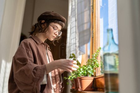 Foto de Niña adolescente cuidando plantas de interior, de pie junto a la ventana en casa revisando hojas y raíces de plantas de interior. Concepto de jardinería y fitosanidad - Imagen libre de derechos