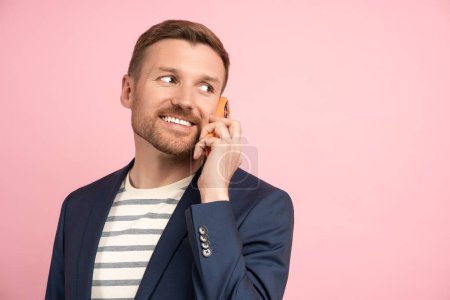 Photo for Smiling businessman talking on mobile phone joyfully looking away isolated on pink background dressed in blue jacket over stylish striped t-shirt. Happy young man solving business questions distantly - Royalty Free Image