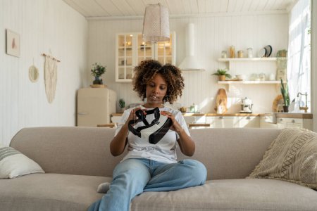 Téléchargez les photos : Femme afro-américaine excitée regardant son premier casque VR et se sentant excitée de se plonger, assise sur un canapé dans le salon. Curieux noir dame gamer tenant futuriste lunettes de réalité virtuelle - en image libre de droit