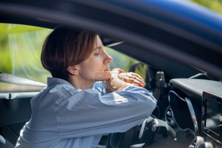 Téléchargez les photos : Pensive femme sérieuse debout dans l'embouteillage attendant voiture de conduite regardant vers l'avenir. Femme calme et réfléchie assise dans une voiture attachée avec la ceinture de sécurité pliant les mains sur le volant. Problèmes routiers urbains. - en image libre de droit