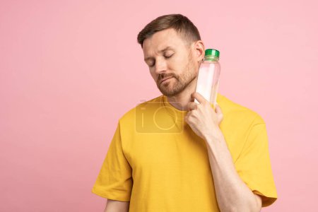 Photo for Overheated tired man suffers from heat, puts bottle of cold water to neck. Exhausted guy trying to cool down after sports training in hot summer day, feels dehydrated and thirsty isolated on pink wall - Royalty Free Image