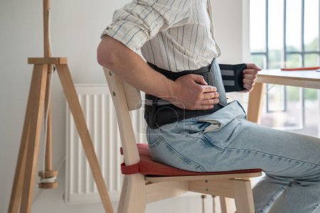 Photo for Man sitting at table wearing medical corset for lumbar spine, suffering from chronic back pain, using spinal decompression belt. Rehabilitation after spinal Injury - Royalty Free Image