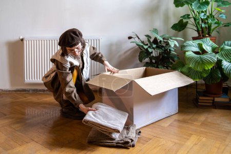 Photo for Clothing donation concept. Millennial girl packing clothes for moving into new apartment, using cardboard box, donating clothing items. Young woman unpacking after move. - Royalty Free Image