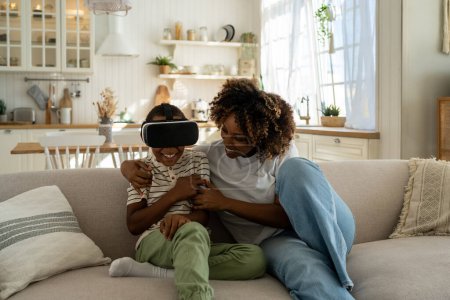 Téléchargez les photos : Heureuse maman afro-américaine et son fils utilisant des lunettes virtuelles pour jouer à des jeux vidéo. Garçon regardant le monde virtuel dans le casque VR assis sur le canapé à la maison. Réalité virtuelle, cyberespace, concept de divertissement. - en image libre de droit