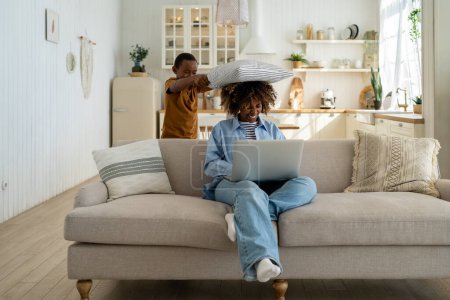 Photo for Pleased playful African American child boy pillow hit to surprised head mom busy work online shopping while sit with laptop on sofa. Happy family time mother and son together fun game activity at home - Royalty Free Image