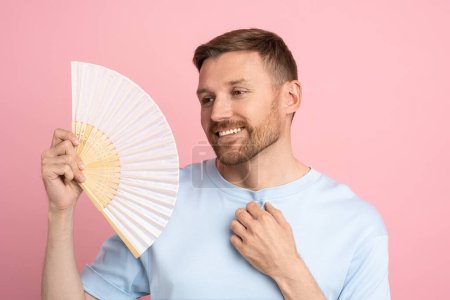 Photo for Relaxed smiling man using paper fan at summer hot weather, sweating, cools himself, enjoying fresh air and closed eyes isolated on studio pink background. Overheating, heat stroke concept. - Royalty Free Image